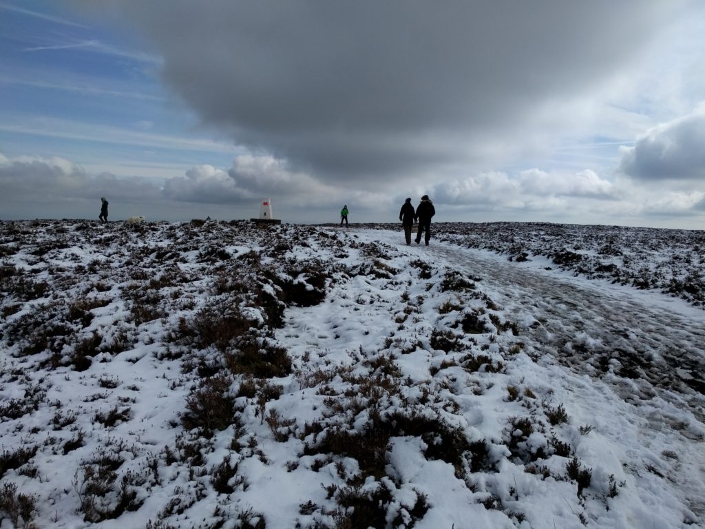People walking on path with a thin layer of snow and trig point in the distance